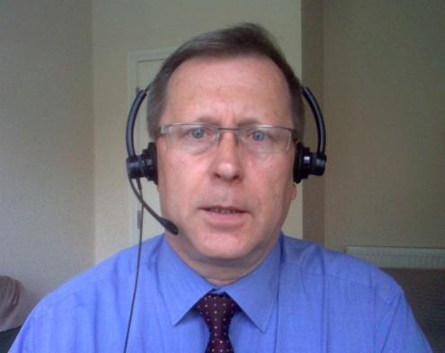 Portret mężczyzny w okularach i niebieskiej koszuli z krawatem w kropki, z zestawem słuchawkowym na głowie, potencjalnie nauczyciel języka angielskiego podczas lekcji online.