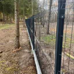 Czarny system ogrodzeniowy z paneli zamontowany na betonowej podmurówce w lesie, widok z boku wzdłuż linii ogrodzenia.