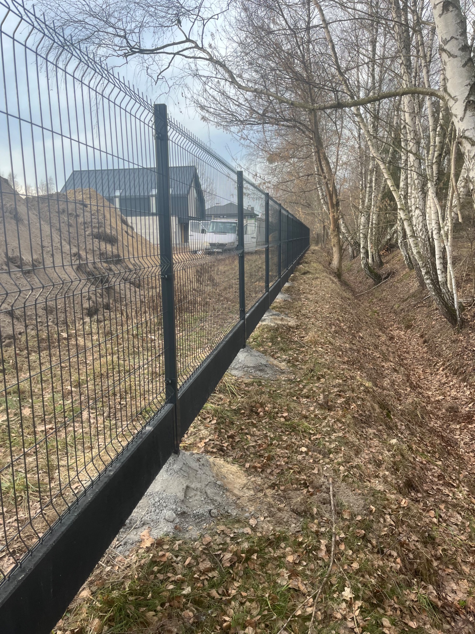 Długie, czarne ogrodzenie panelowe z widocznymi fundamentami z betonu, biegnące wzdłuż rowu z opadłymi liśćmi, w tle dom i biały samochód dostawczy