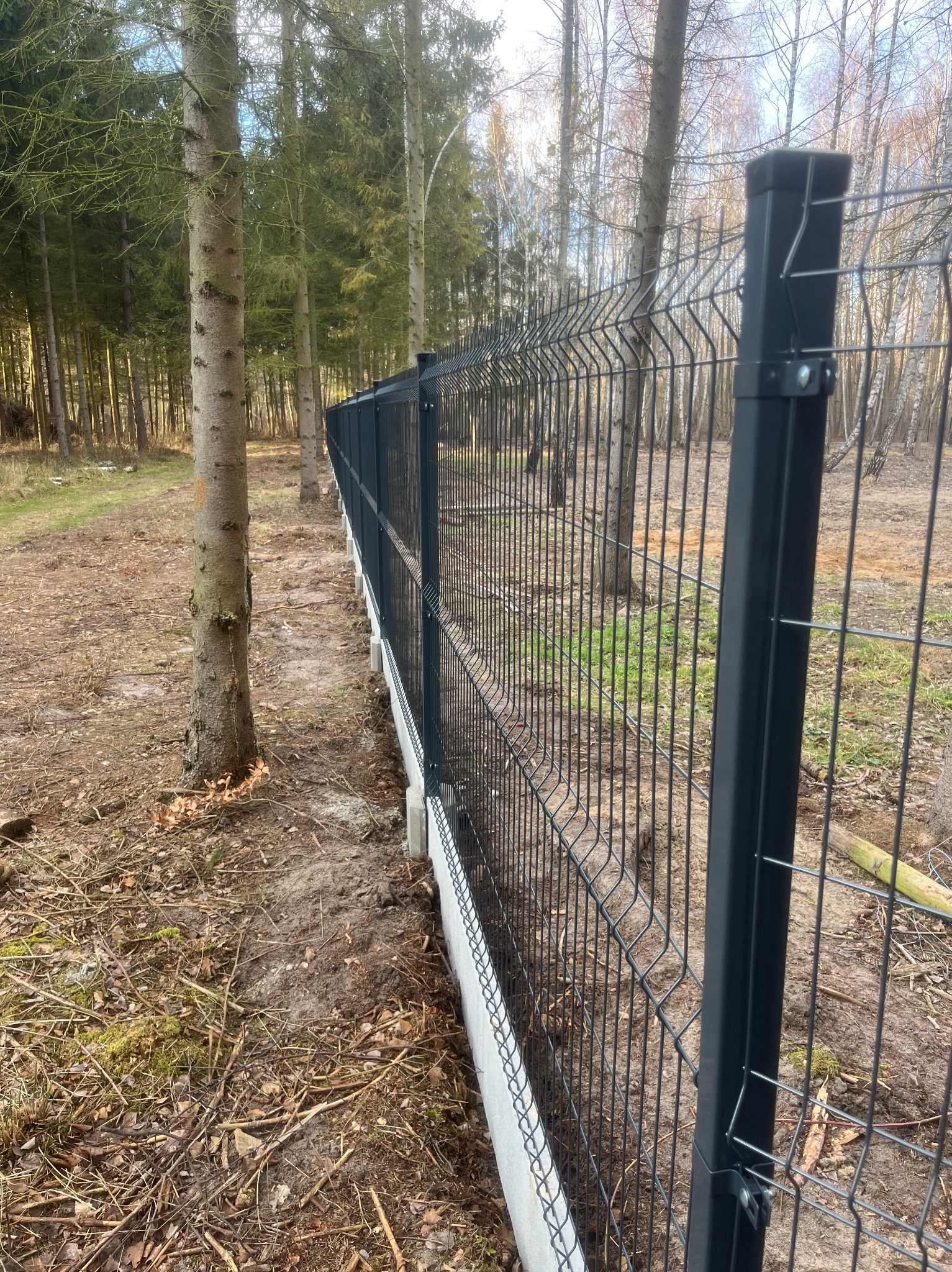 Czarny system ogrodzeniowy z paneli zamontowany na betonowej podmurówce w lesie, widok z boku wzdłuż linii ogrodzenia.