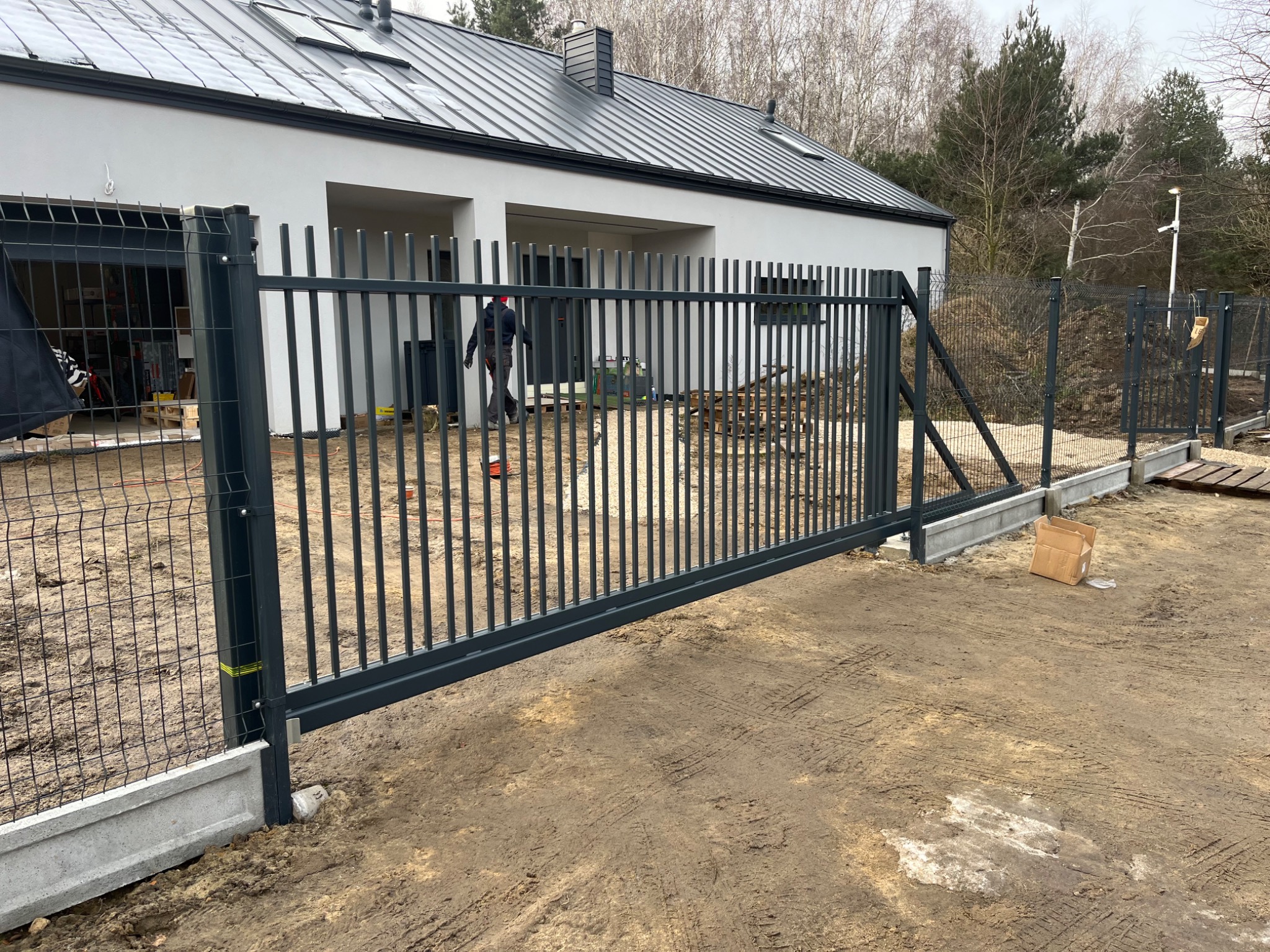 Nowoczesna posesja z przesuwną bramą metalową w kolorze grafitowym, widoczna podczas montażu, z fragmentem ogrodzenia panelowego w tle i pracownikiem w oddali.