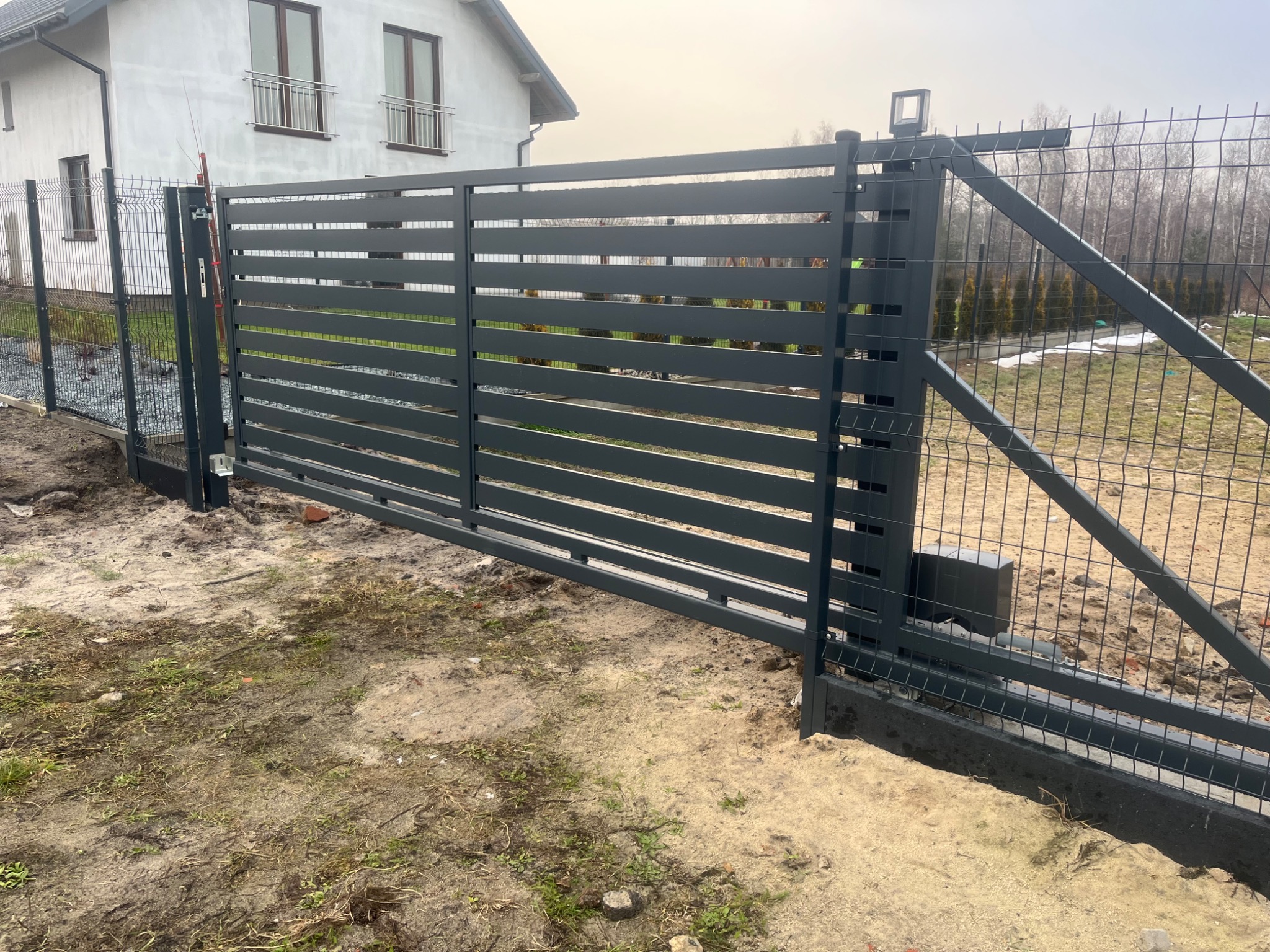 Nowoczesna, przesuwna brama wjazdowa z poziomymi panelami, zintegrowana z ogrodzeniem z siatki, widoczny napęd elektryczny, tło stanowi biały dom i drzewa bez liści.