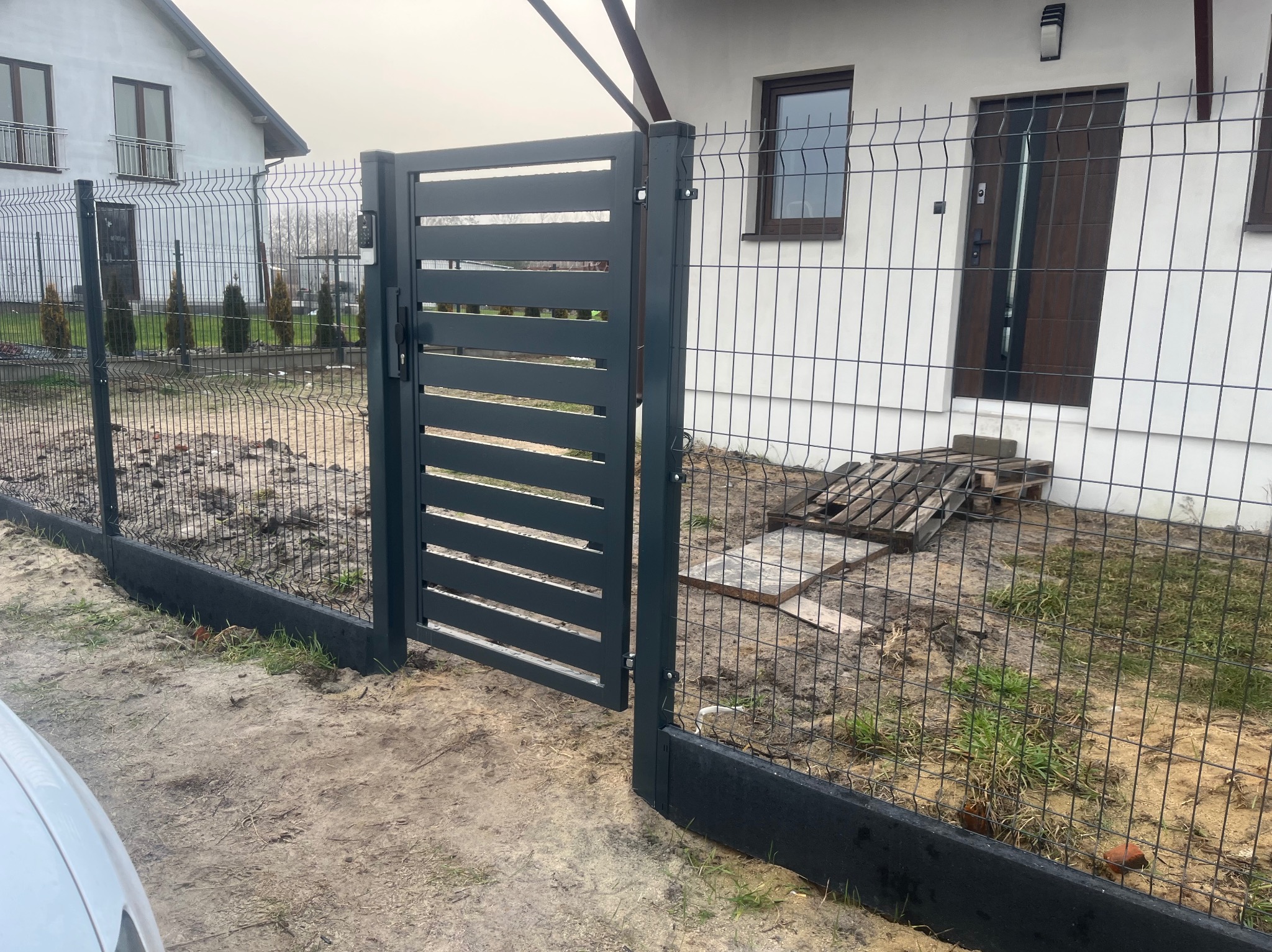 Nowoczesna, grafitowa furtka z poziomych paneli, wkomponowana w siatkę ogrodzeniową z podmurówką, na tle domu z białymi ścianami i brązowymi oknami, widoczne palety za ogrodzeniem.