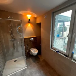 Nowoczesna łazienka z kabiną prysznicową typu walk-in, wiszącą toaletą z bidetką, oświetleniem punktowym i dużym oknem z widokiem na zewnątrz. Na parapecie okna widoczne narzędzia i materiały...