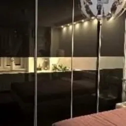 Czarna szafa przesuwna z lustrzanymi drzwiami, przedzielona poziomym pasem. Widoczne oświetlenie LED wewnątrz szafy i dekoracyjna lampa sufitowa. Ujęcie z boku łóżka z fioletową narzutą.