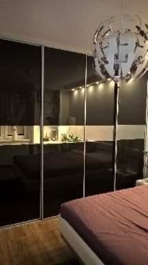 Czarna szafa przesuwna z lustrzanymi drzwiami, przedzielona poziomym pasem. Widoczne oświetlenie LED wewnątrz szafy i dekoracyjna lampa sufitowa. Ujęcie z boku łóżka z fioletową narzutą.