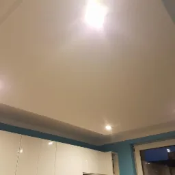 Gładka, jasna powierzchnia sufitu napinanego z trzema punktami świetlnymi LED, widok od dołu, fragment białych szafek kuchennych i okno z widokiem na nocne niebo.