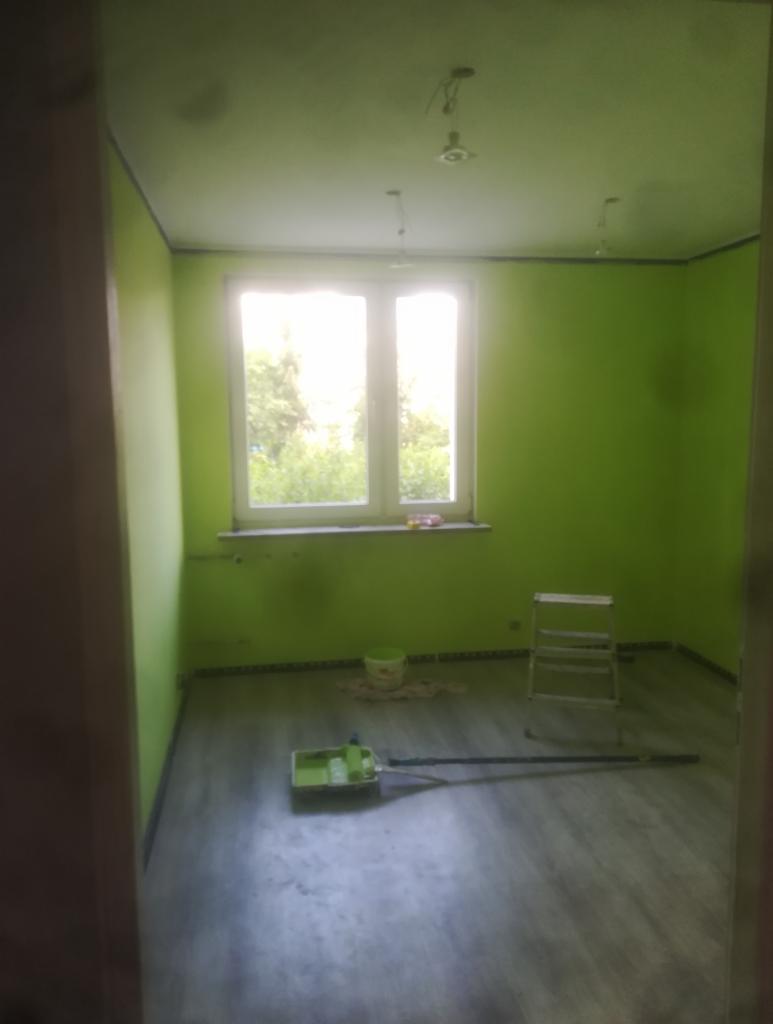 Pokój po świeżym pomalowaniu na jaskrawozielony kolor; widoczne narzędzia malarskie, drabina i niedokładności przy listwach przypodłogowych.
