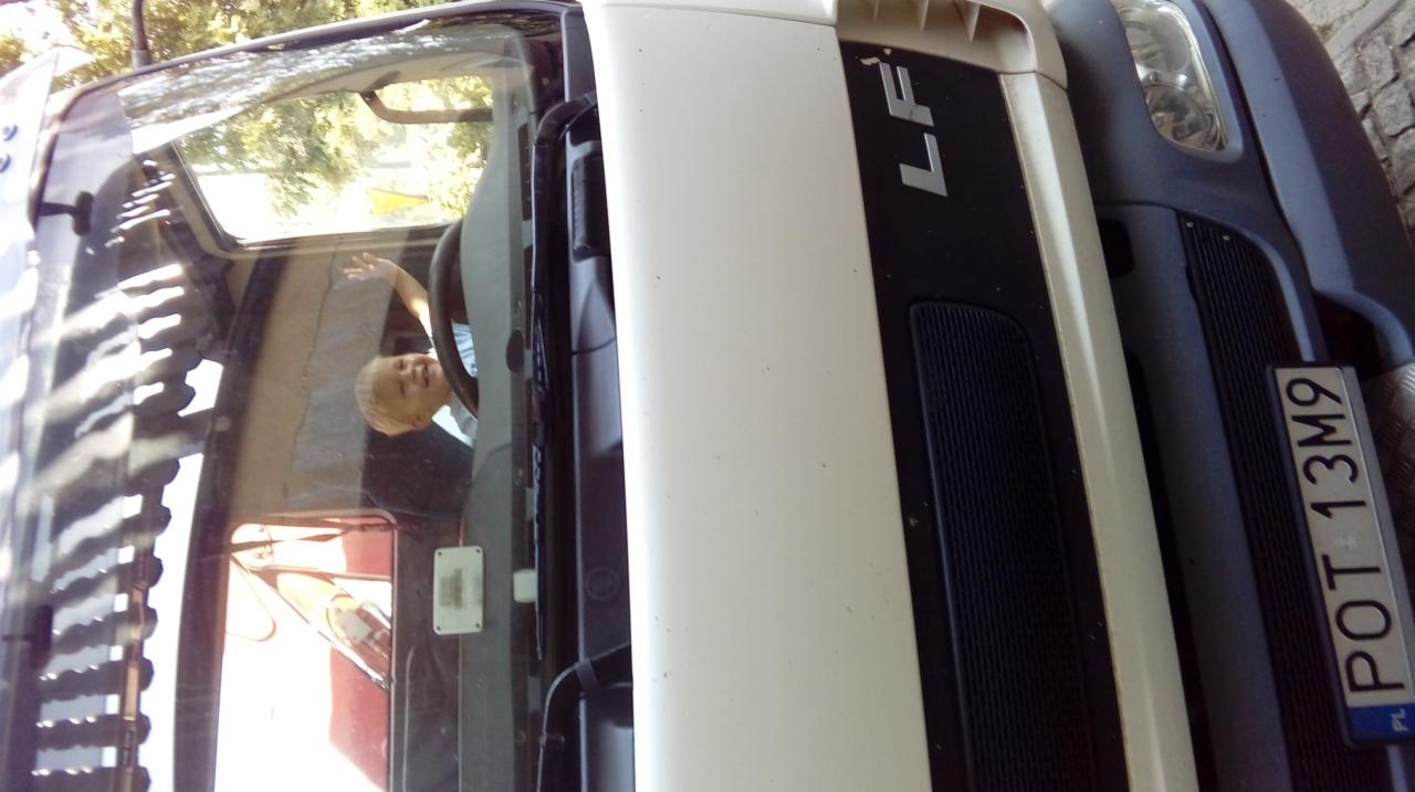 Uśmiechnięte dziecko macha zza szyby białego busa marki LF z widoczną tablicą rejestracyjną.