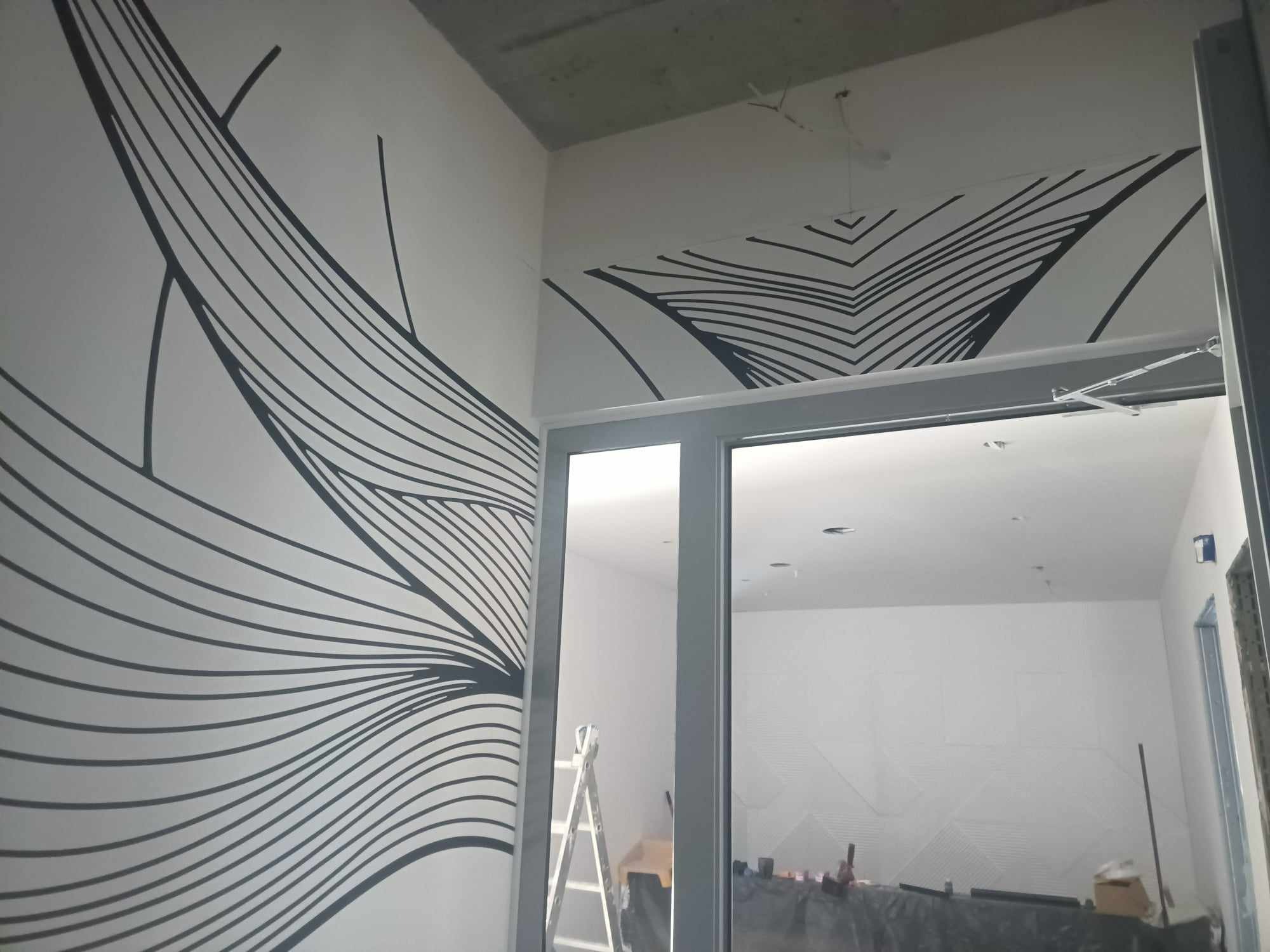 Geometryczny wzór z czarnych linii na białej ścianie, częściowo widoczny nad drzwiami, z surowym betonowym sufitem i drabiną w tle.