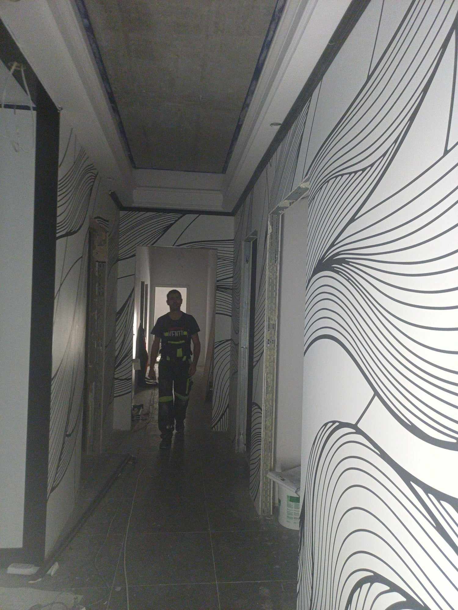 Wnętrze korytarza w trakcie remontu, ściany pokryte tapetą w czarno-białe, abstrakcyjne wzory, widoczny pracownik w oddali, surowy sufit i framugi drzwi.