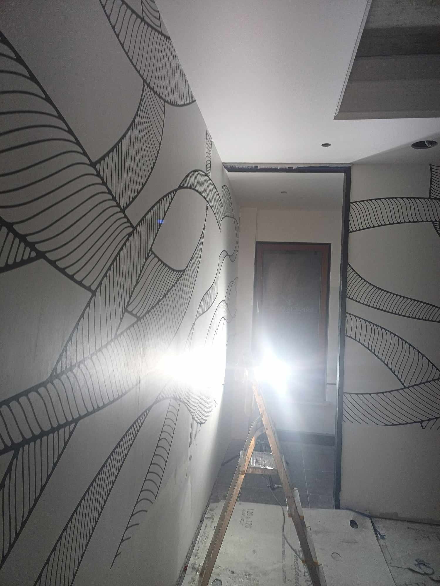 Ściana z dekoracyjnym, czarnym wzorem liści na szarym tle, widoczna drabina i fragment sufitu z oświetleniem. W tle otwarte drzwi z widocznym pomieszczeniem.