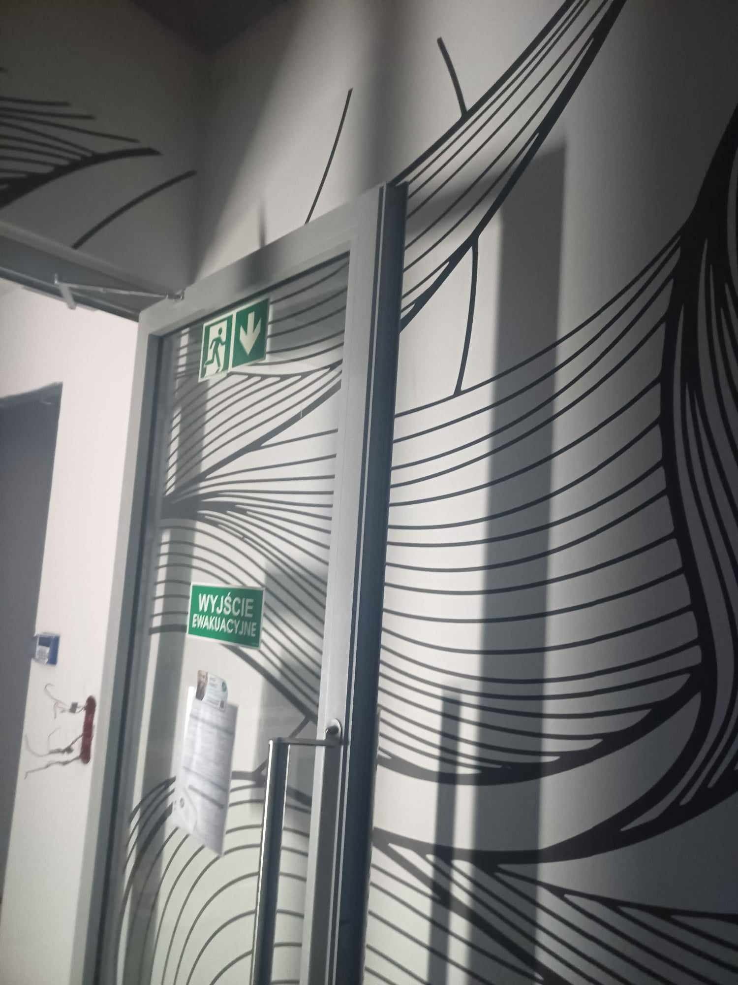 Biała ściana z czarnym, abstrakcyjnym wzorem przypominającym fale, częściowo zasłonięta przez szklane drzwi z napisem 'Wyjście Ewakuacyjne' i symbolem kierunku ewakuacji. Na drzwiach przyklejone...