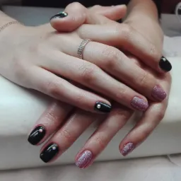 Dłonie kobiety z różnymi stylizacjami paznokci: czarne z pojedynczym kryształkiem i brokatowe w odcieniach różu, na serdecznym palcu widoczny pierścionek z drobnymi kamieniami, na nadgarstku...