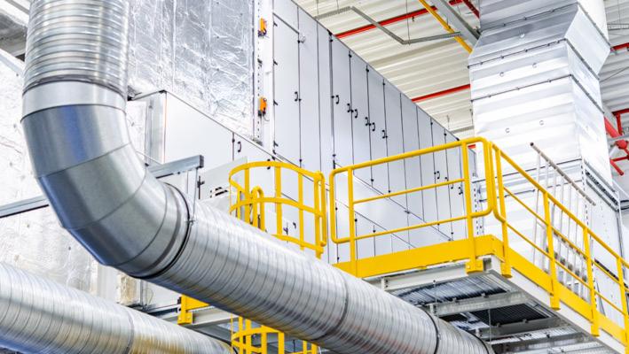 System wentylacyjny z metalowymi rurami spiralnymi i żółtymi barierkami ochronnymi na tle białych paneli w hali przemysłowej.