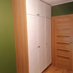 Wysoka szafa wnękowa z drewnianymi bokami i białymi frontami z delikatnym, falistym wzorem, dopasowana do koloru drewnianej podłogi i drzwi, na tle oliwkowozielonej ściany.