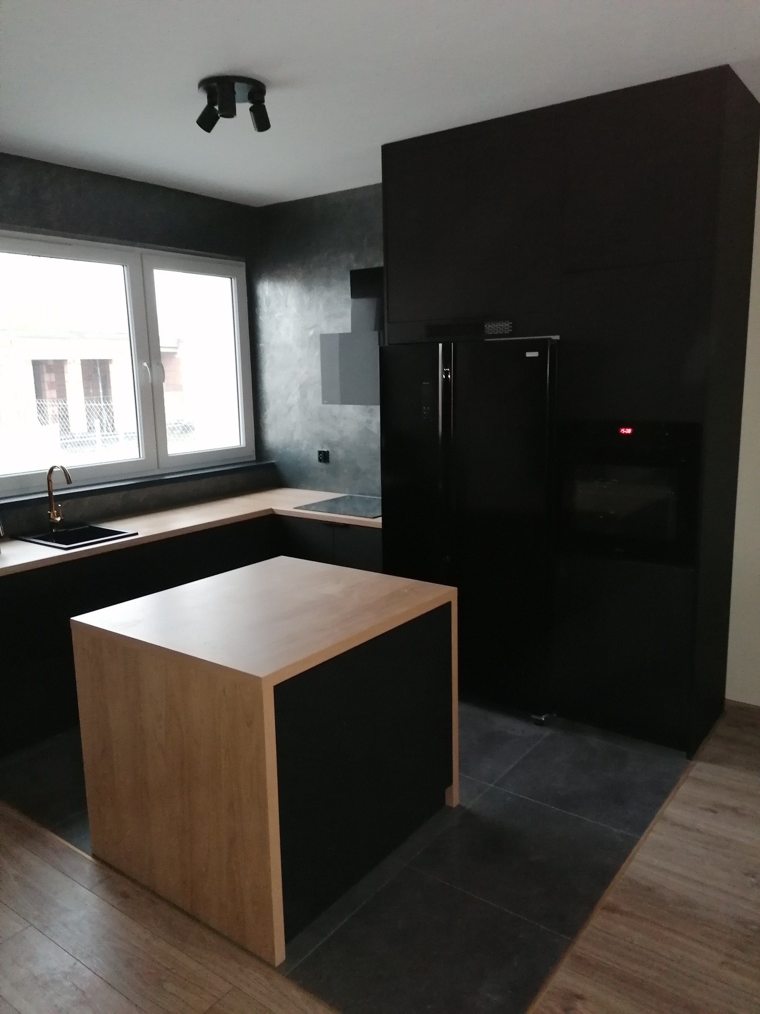 Nowoczesna kuchnia w odcieniach szarości i czerni z drewnianym blatem wyspy, minimalistycznym oświetleniem sufitowym i złotą armaturą przy zlewie.