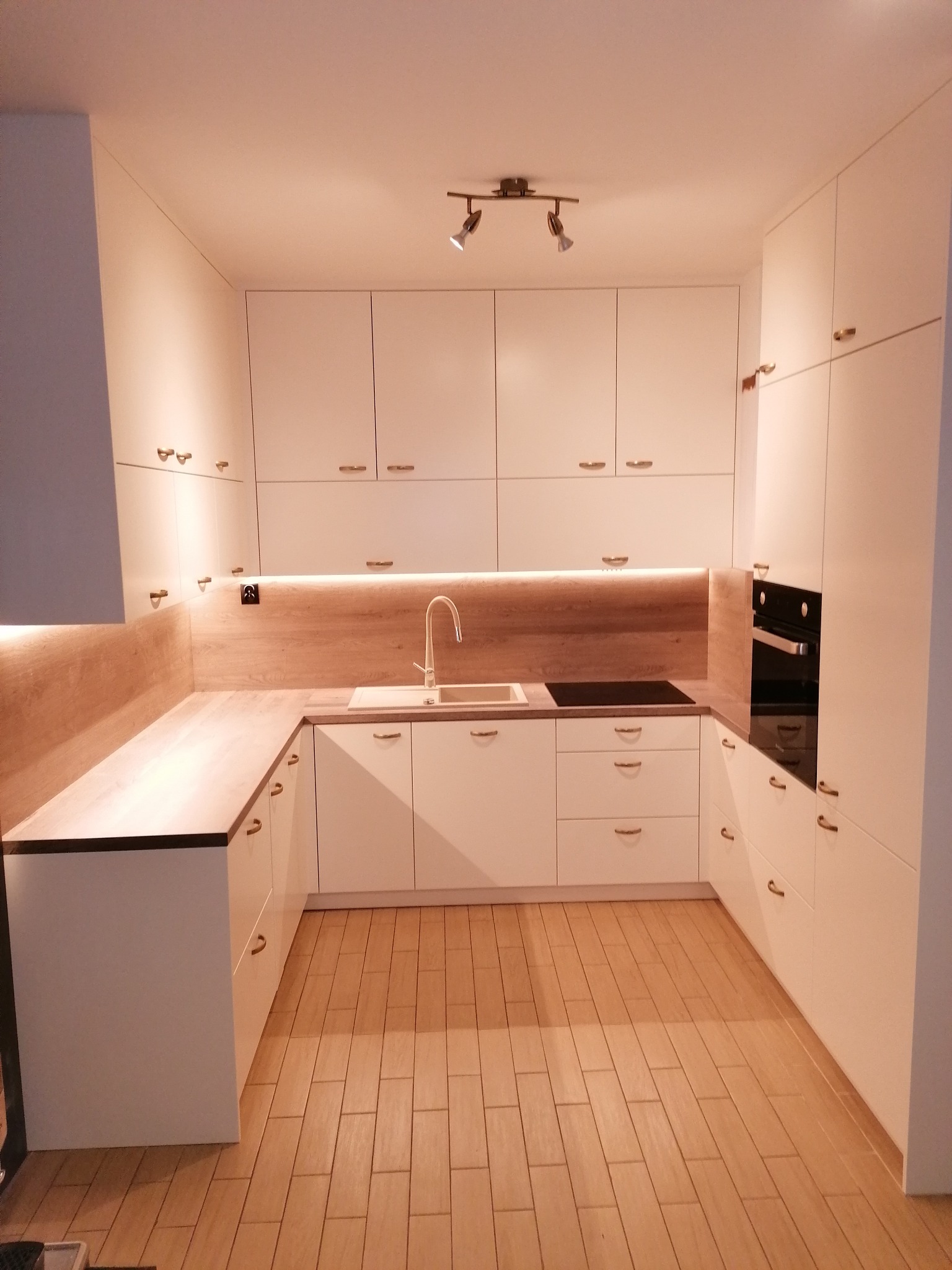 Nowoczesna, jasna kuchnia z białymi frontami, drewnianym blatem i podświetleniem LED nad blatem, złote uchwyty, piekarnik w zabudowie i podłoga imitująca drewno.