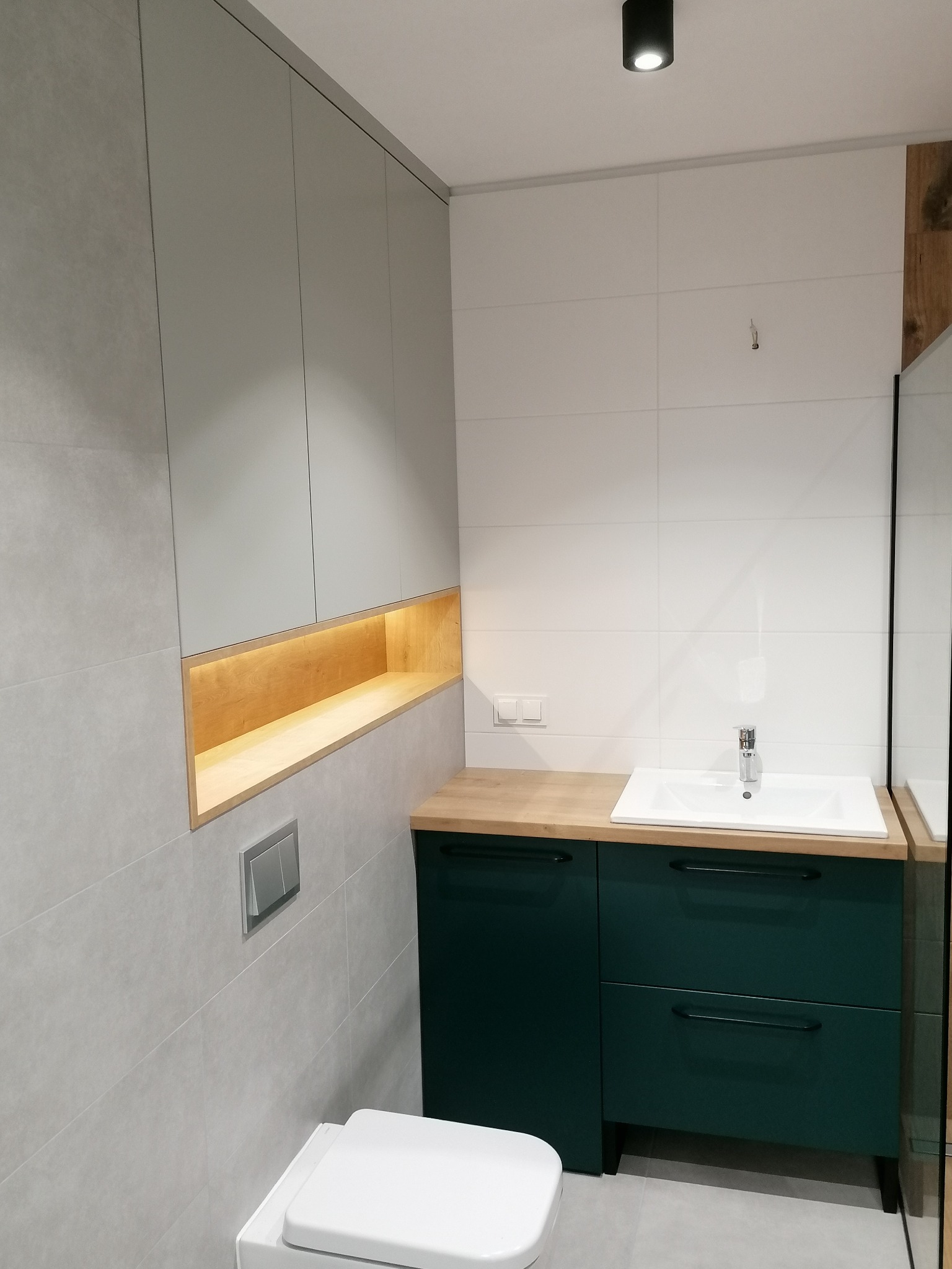 Nowoczesna łazienka z szarymi szafkami wiszącymi z podświetlaną wnęką, zieloną szafką pod umywalką z drewnianym blatem i białą ceramiką sanitarną na szarych płytkach.