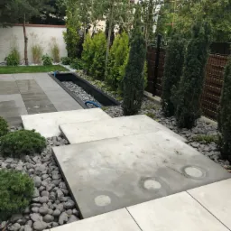 Nowoczesny ogród z betonowymi płytami przejściowymi, szarą kostką dekoracyjną, wąskim zbiornikiem wodnym z niebieskim wężem, tujami i innymi roślinami ozdobnymi.