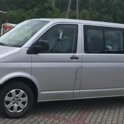 Srebrny Volkswagen Transporter T5 zaparkowany na brukowanym placu, widok z boku na tle zieleni.