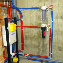 ECOLIFE Sp. z o.o. - Instalacja wodno-kanalizacyjna w surowym stanie: widoczne rury w kolorach czerwonym i niebieskim, filtry wody, stelaż podtynkowy WC marki Wisa, ściana z cegły i surowy beton
