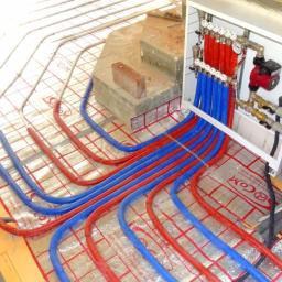 ECOLIFE Sp. z o.o. - Instalacja ogrzewania podłogowego w trakcie montażu: widoczne rury grzewcze w kolorach czerwonym i niebieskim ułożone na folii izolacyjnej z nadrukiem, rozdzielacz zamontowany na ścianie z cegły.