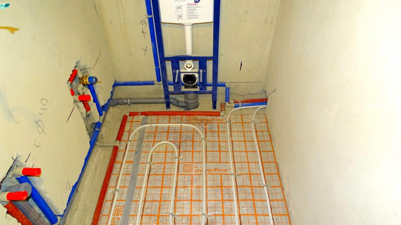 Wnętrze łazienki w trakcie montażu instalacji ogrzewania podłogowego i podwieszanego WC, widoczne rury grzewcze na pomarańczowej macie, nieotynkowane ściany z wystającymi elementami instalacji wodnej.