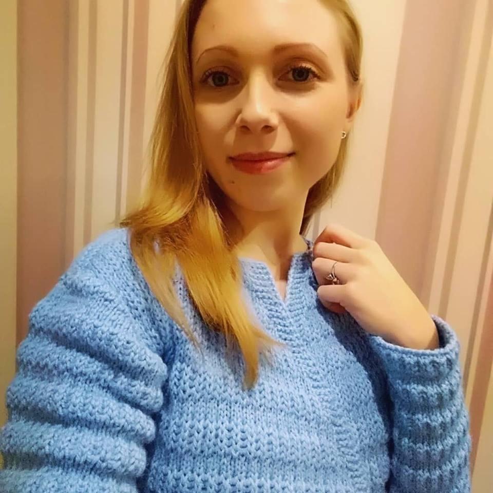 Portret kobiety w błękitnym, ręcznie robionym swetrze z wyraźnym splotem, na tle ściany w pionowe, pastelowe pasy. Kobieta ma blond włosy i delikatny makijaż.