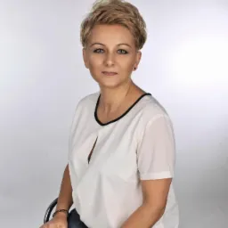 Portret kobiety z krótkimi blond włosami, ubranej w białą bluzkę z czarną lamówką, siedzącej na krześle na białym tle. Na nadgarstku złoty zegarek.