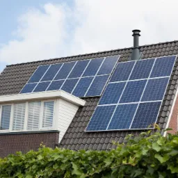 Panele słoneczne na dwuspadowym dachu domu z cegły i dachówki, widok z poziomu gruntu z zielonym żywopłotem w dolnej części kadru.