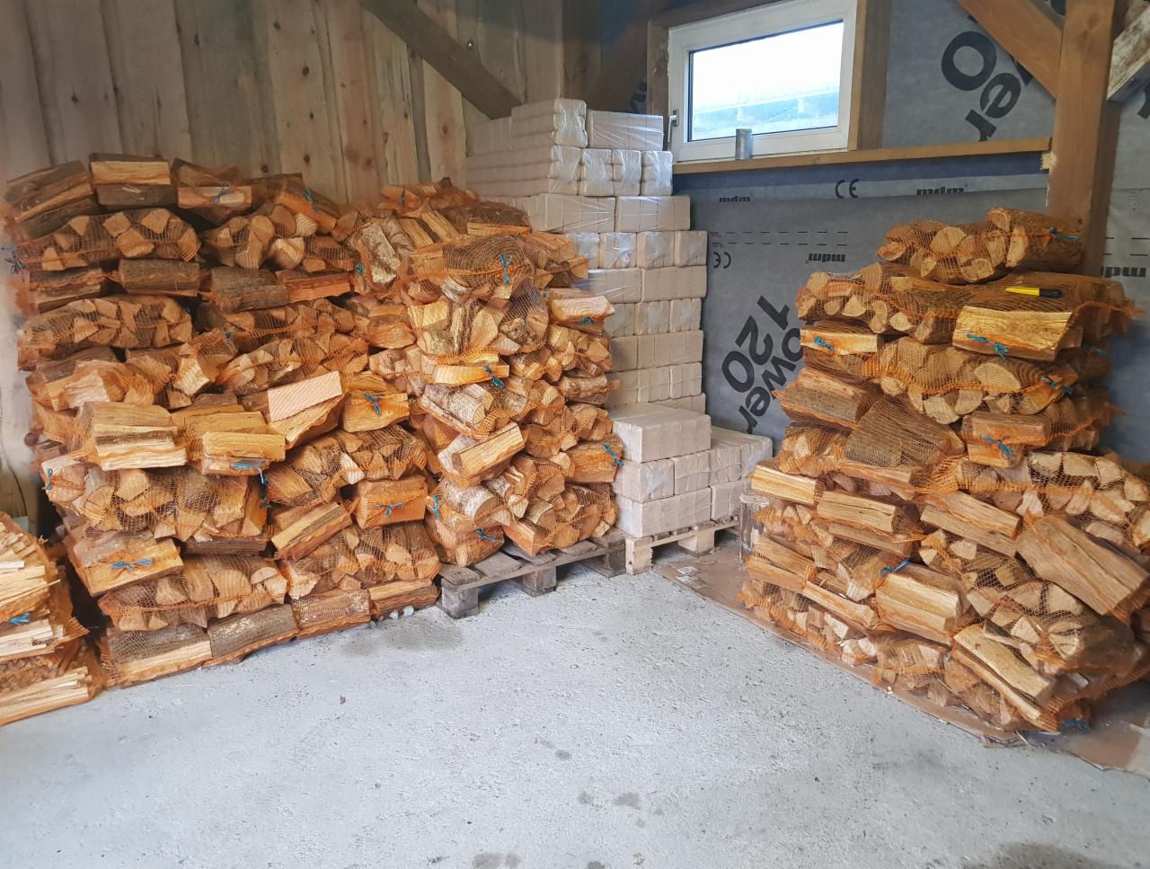 Skladowane worki z pociętym drewnem na opał i stosy brykietu drzewnego w magazynie, widoczne drewniane ściany i okno.