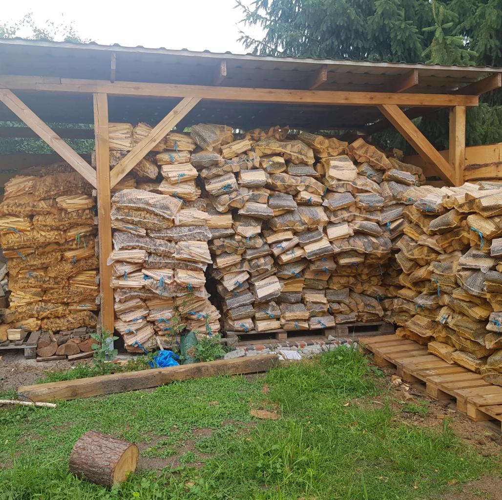 Zadaszony skład z paletami pełnymi workowanego drewna opałowego, widok z poziomu trawy.