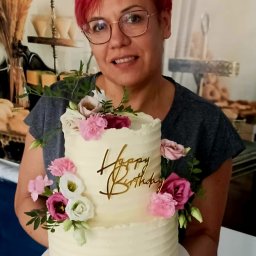 Ciacha Ewy Pracownia Tortów Artystycznych Zielona Góra - Kobieta z różowymi włosami prezentuje dwupiętrowy tort urodzinowy z kremem i kwiatami w Zielonej Górze. Dekoracja z napisem 'Happy Birthday'.