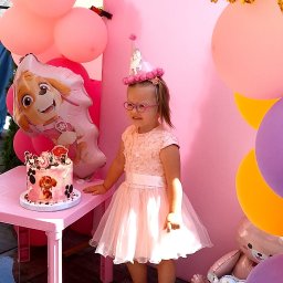 Ciacha Ewy Pracownia Tortów Artystycznych Zielona Góra - Uśmiechnięta dziewczynka w różowej sukience i czapeczce urodzinowej stoi obok tortu Paw Patrol na tle różowych balonów i dekoracji urodzinowych.