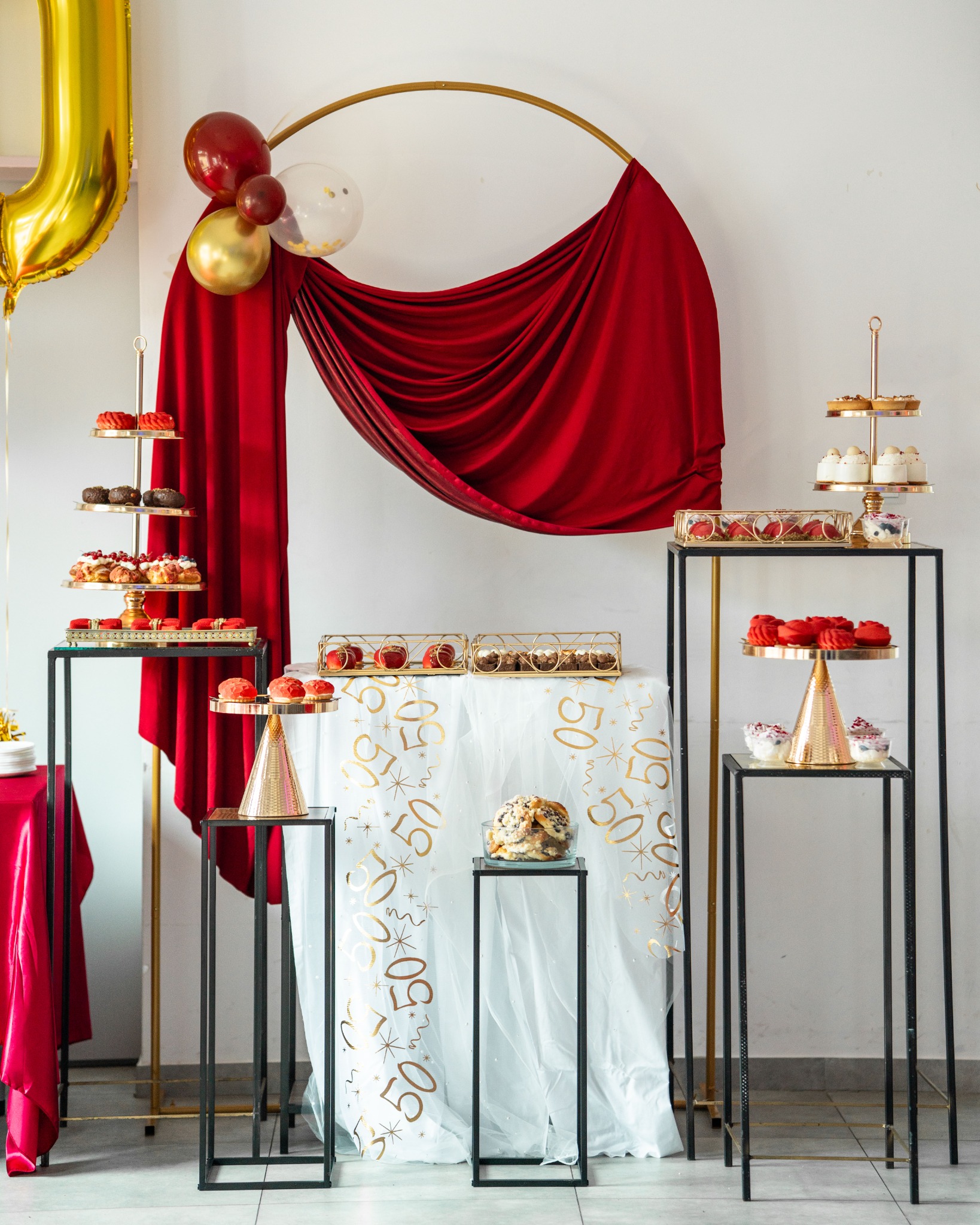 Elegancki stół z tortami, babeczkami i dekoracjami na 50 urodziny. Czerwone draperie, złote balony i biały obrus z motywem cyfry 50.