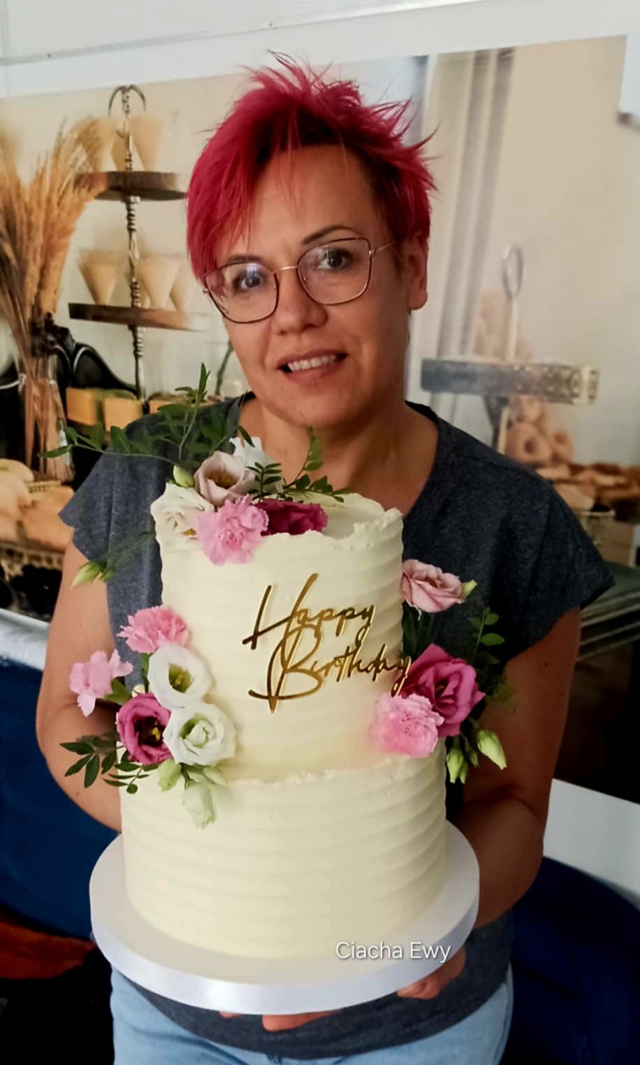 Kobieta z różowymi włosami prezentuje dwupiętrowy tort urodzinowy z kremem i kwiatami w Zielonej Górze. Dekoracja z napisem 'Happy Birthday'.