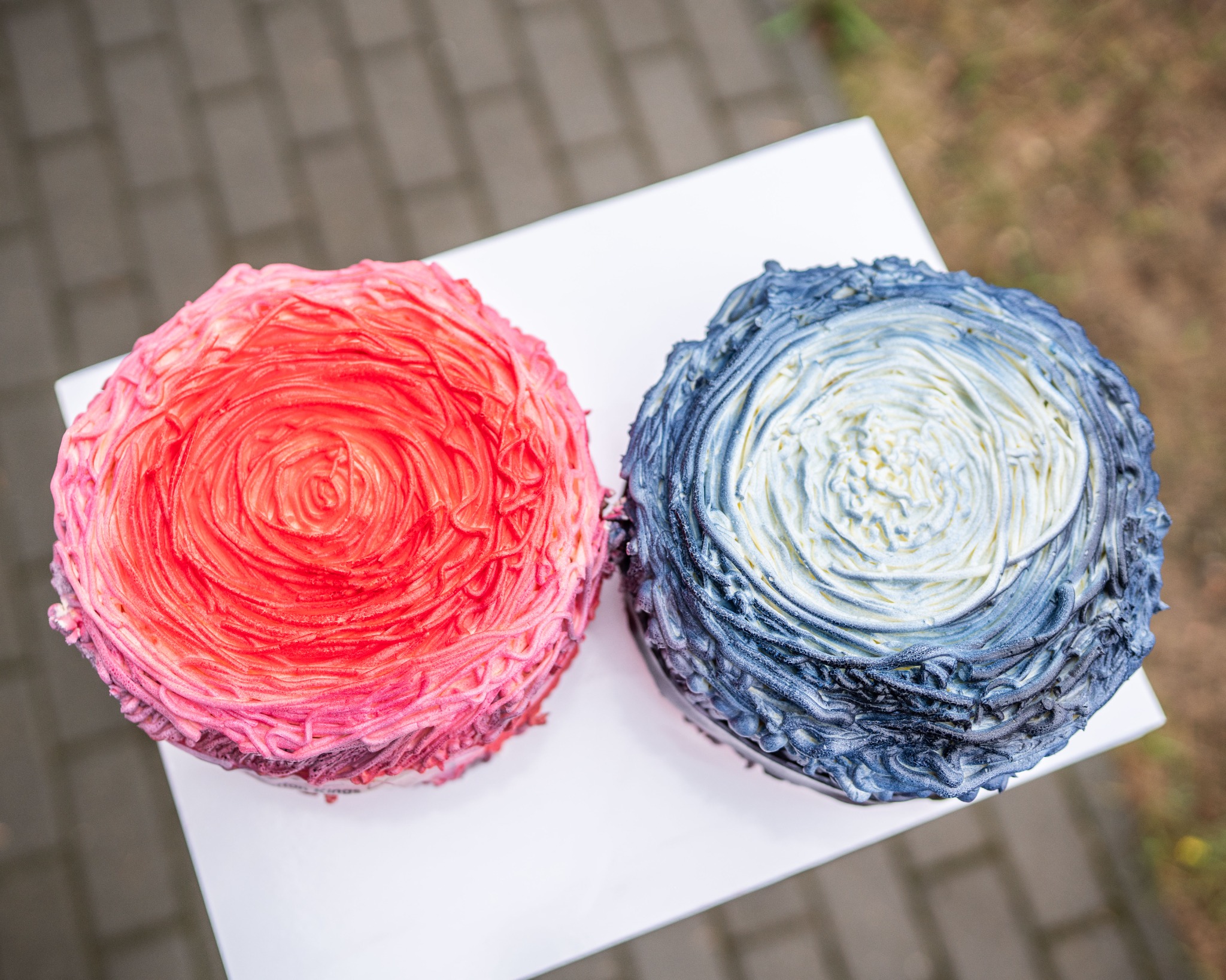 Dwa artystyczne torty: różowy z kremem w kształcie róży i niebieski z kremem imitującym fale, na białym podeście, ukazane z góry.