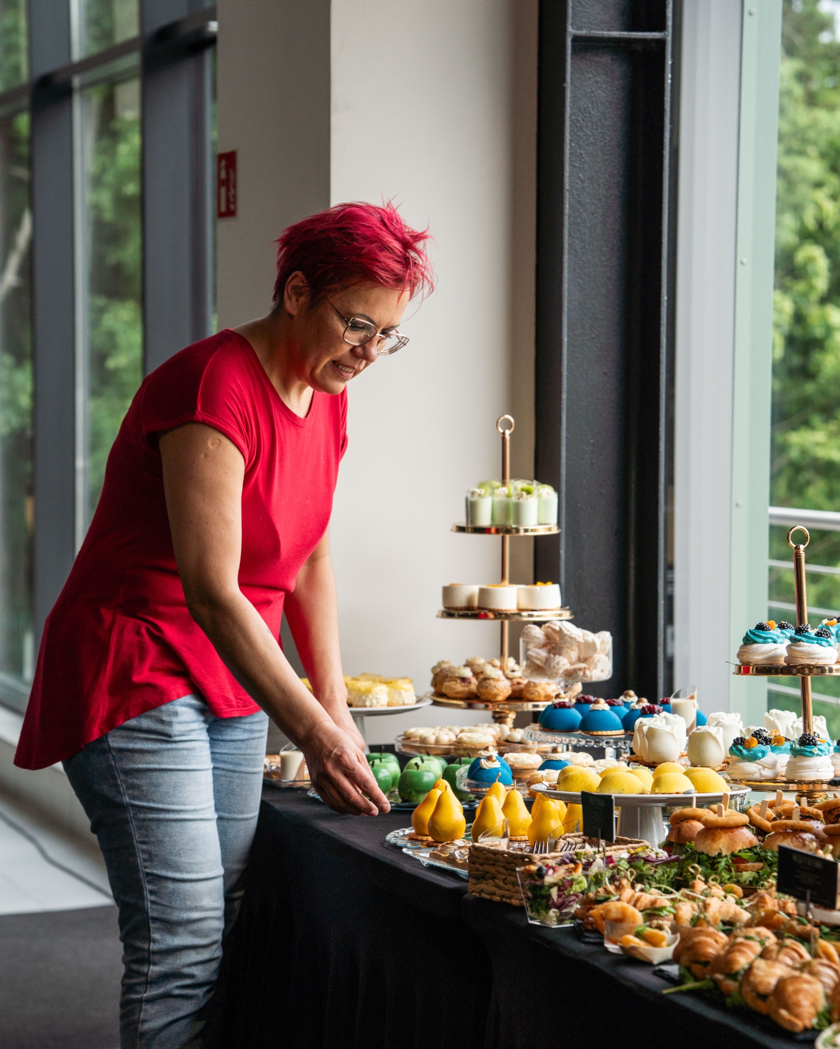Kobieta w czerwonej koszulce układa różnorodne desery i przekąski na stole podczas wydarzenia. W tle duże okno z widokiem na zieleń.