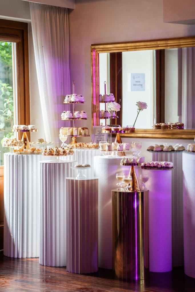 Elegancki bufet słodkości na białych i złotych kolumnach, oświetlony fioletowym światłem, z lustrem w tle. Różnorodne ciastka i desery na paterach.
