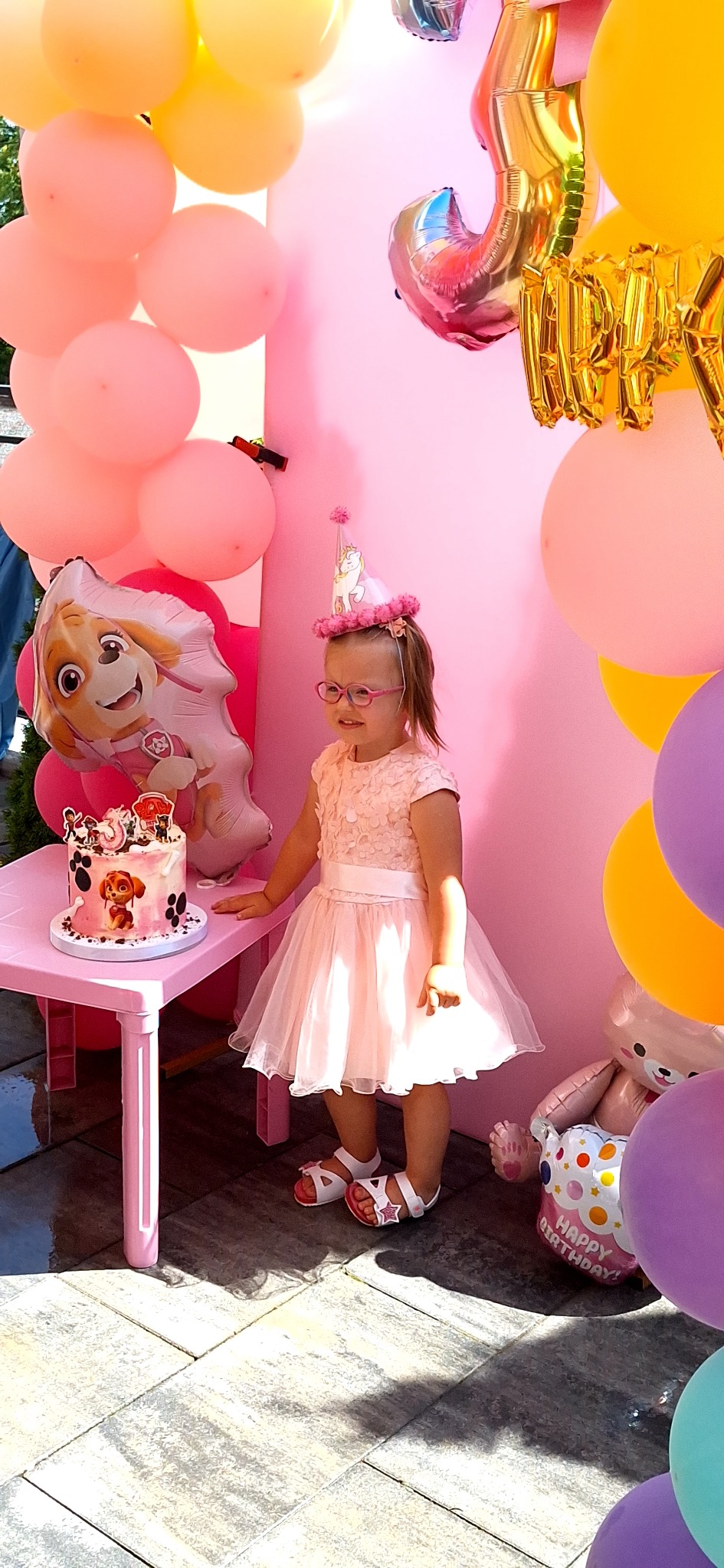 Uśmiechnięta dziewczynka w różowej sukience i czapeczce urodzinowej stoi obok tortu Paw Patrol na tle różowych balonów i dekoracji urodzinowych.