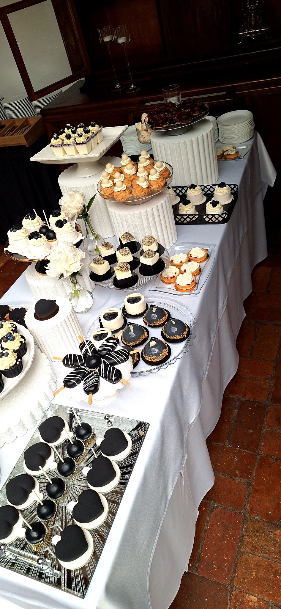 Elegancki stół z deserami w odcieniach bieli i czerni: babeczki z borówkami, ciastka, cake pops i inne słodkości. Dekoracje kwiatowe i szkło dodają szyku.