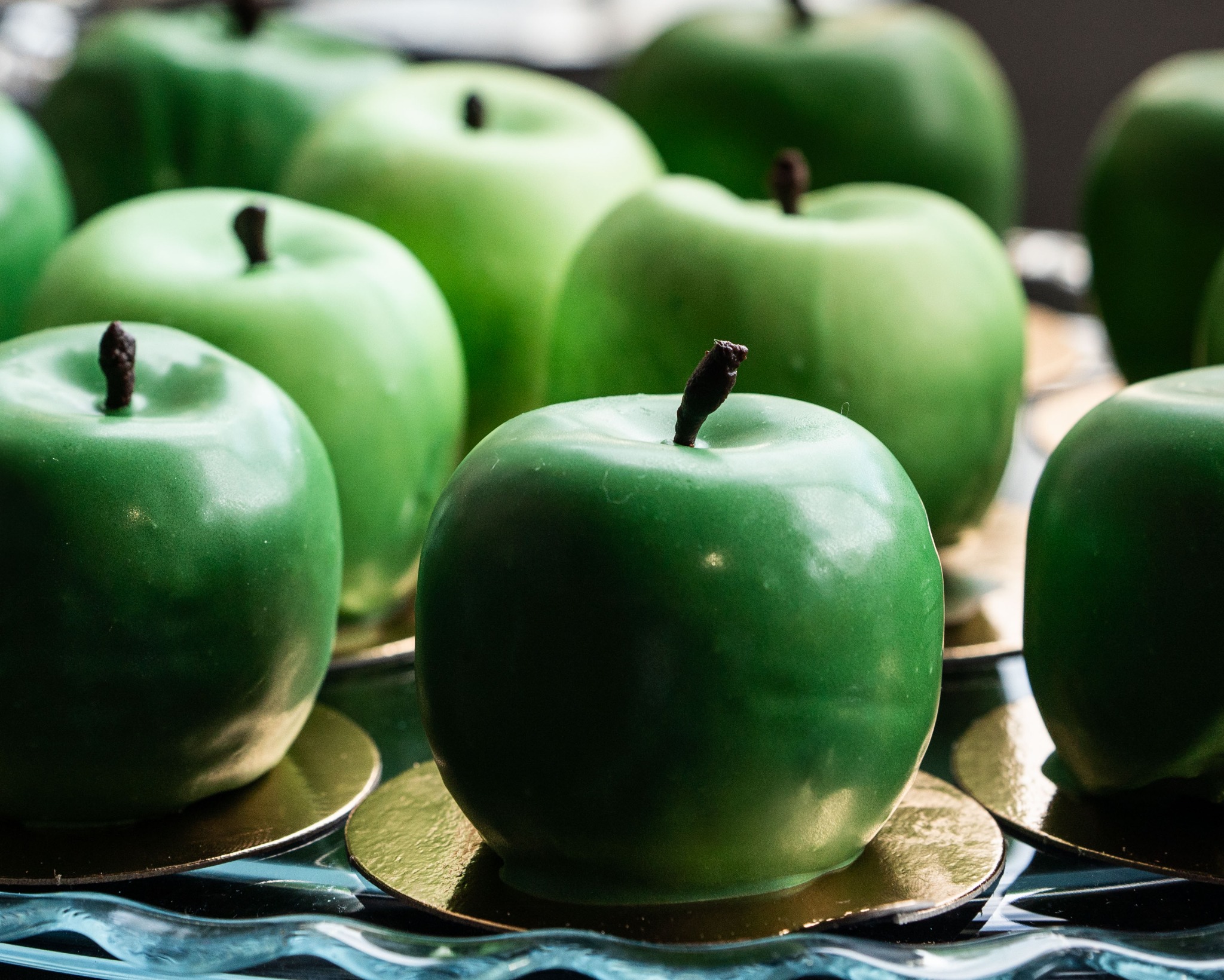 Zielone jabłka z czekolady na złotych podkładkach, ułożone na szklanej paterze. Dekoracyjne słodycze, zbliżenie, gładka powierzchnia.