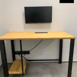 Biurko z blatem z jasnego drewna i czarnymi metalowymi nogami, z dodatkową półką pod blatem, na ścianie zamontowany monitor i logo firmy.