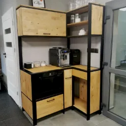 Kuchenna zabudowa narożna z jasnego drewna i czarnych metalowych ram, z wbudowaną zmywarką i ekspresem do kawy, uzupełniona dekoracyjnymi butelkami na półce i plakatem 'But first coffee'.