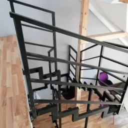 Nowoczesne schody o konstrukcji metalowej z czarnymi, geometrycznymi elementami, widok z góry, drewniane podłogi i elementy konstrukcyjne dachu, krzesła w odcieniach fioletu w tle.