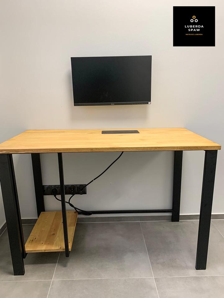 Biurko z blatem z jasnego drewna i czarnymi metalowymi nogami, z dodatkową półką pod blatem, na ścianie zamontowany monitor i logo firmy.