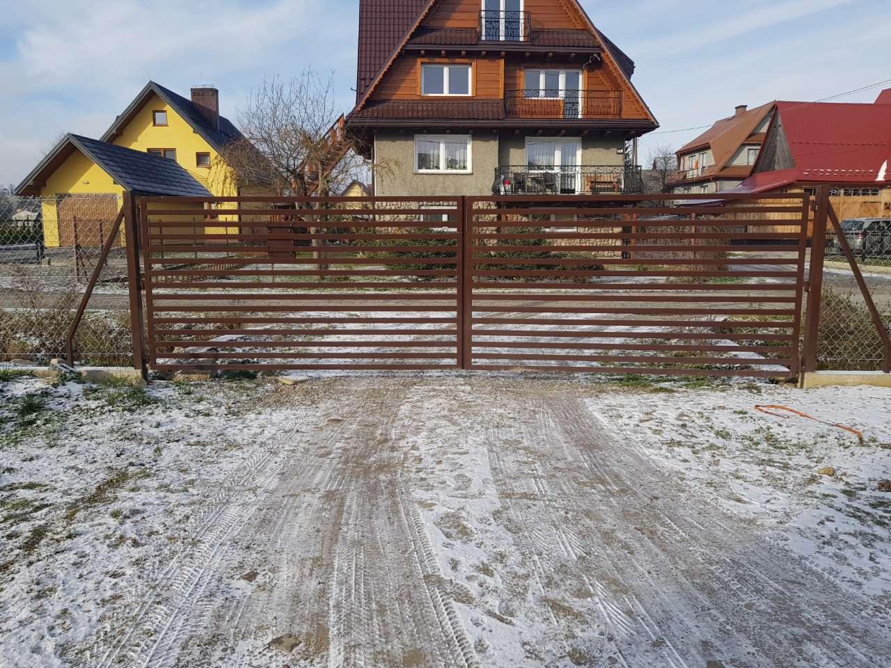 Brązowa brama posesyjna z poziomych profili stalowych na tle zabudowy jednorodzinnej w zimowej scenerii, widoczne ślady opon na śniegu.