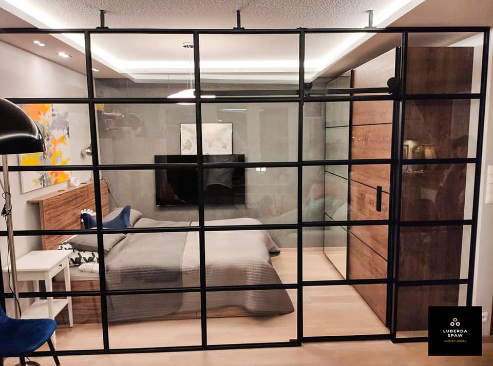 Przeszklona ścianka działowa z czarnymi profilami oddzielająca sypialnię od reszty mieszkania, widoczna szafa przesuwna z drewnianymi frontami i łóżko z szarą pościelą.