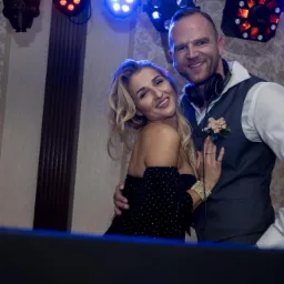 Uśmiechnięta para, kobieta w czarnej sukience i mężczyzna w kamizelce z butonierką i słuchawkami na szyi, pozują na tle wzorzystej ściany i oświetlenia eventowego.